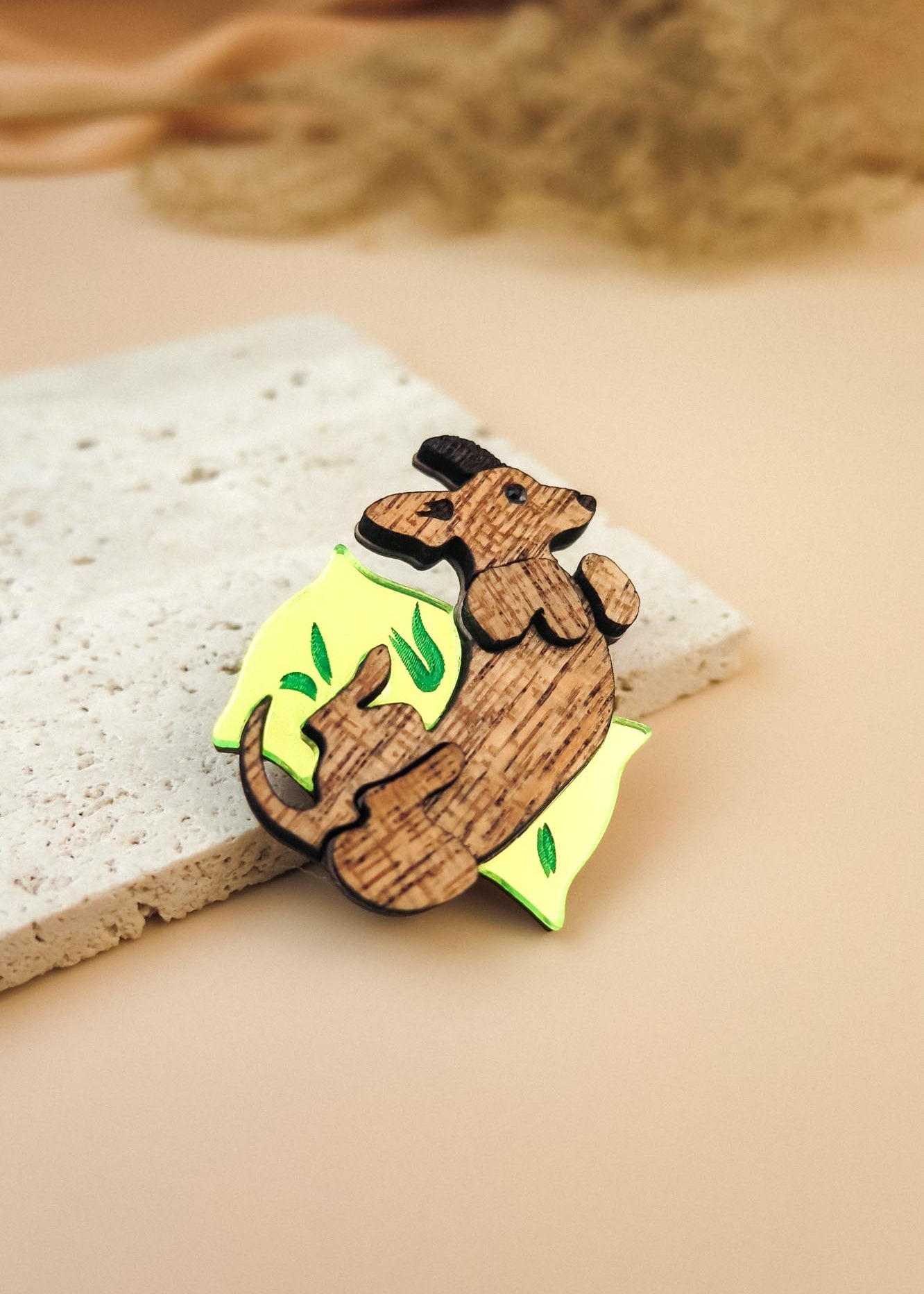 A playful brooch, this wooden pin shaped like a dachshund lounging on a yellow wedge with green leaf accents rests on textured beige stone, making it a charming dog accessory or a unique Dachshund brooch for any outfit.