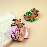 Two wooden pins shaped like dachshunds, one lying on a pink pillow with leaf designs and the other on a green pillow. Both are displayed as playful jewelry on a light beige surface next to a textured stone.