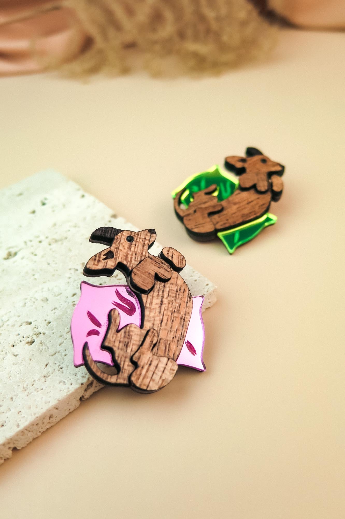 Two wooden pins shaped like dachshunds, one lying on a pink pillow with leaf designs and the other on a green pillow. Both are displayed as playful jewelry on a light beige surface next to a textured stone.