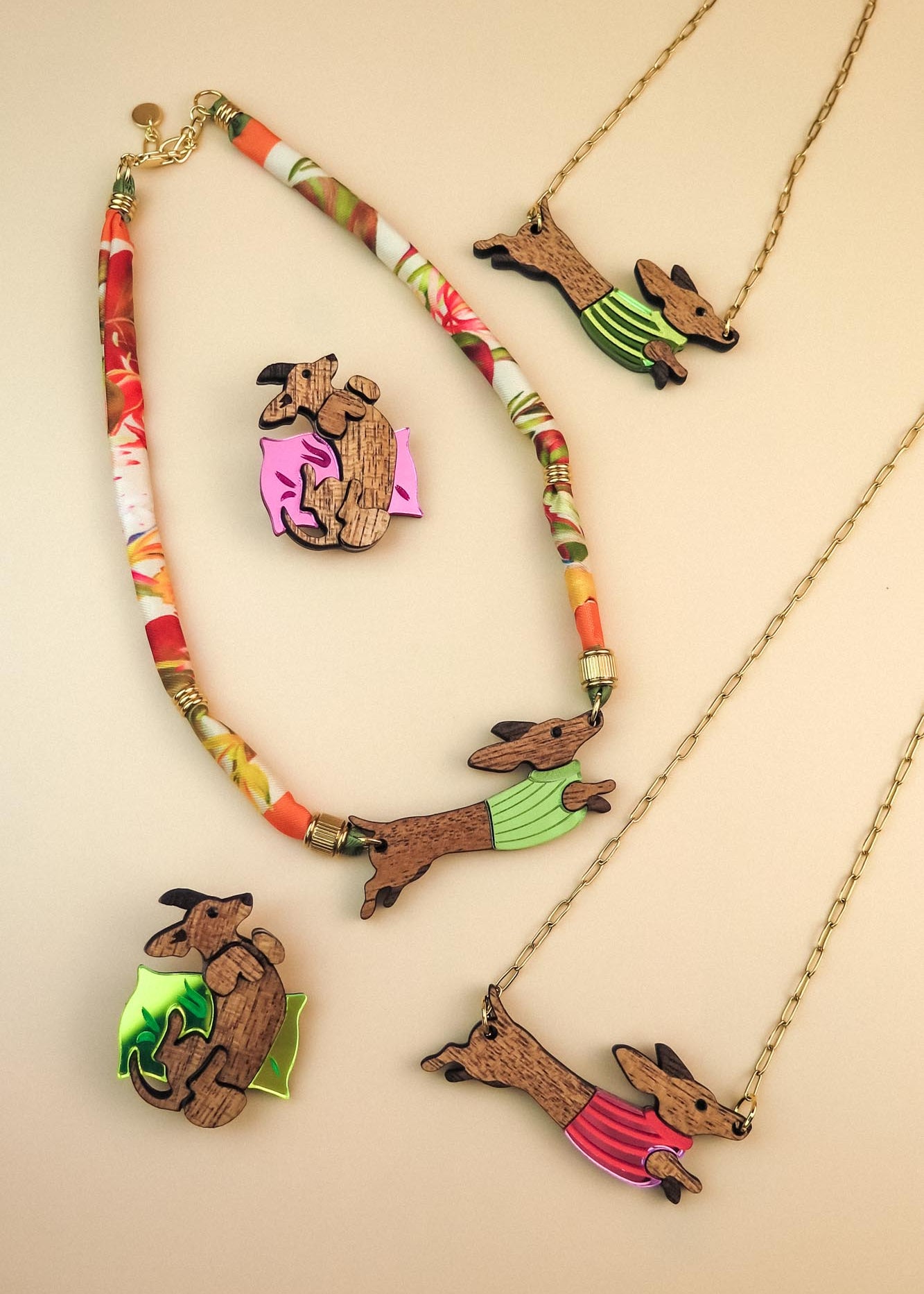Colorful jewelry set featuring dachshund-shaped necklaces and brooches; the dogs wear green or pink colorful sweaters, with beads and gold chain accents, crafted from sustainable wood and arranged on a neutral beige background.