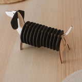 A napkin holder shaped like a dachshund dog, made of wood and black felt, sits on a light wooden table with woven placemats and matching Dachshund coasters visible in the background.