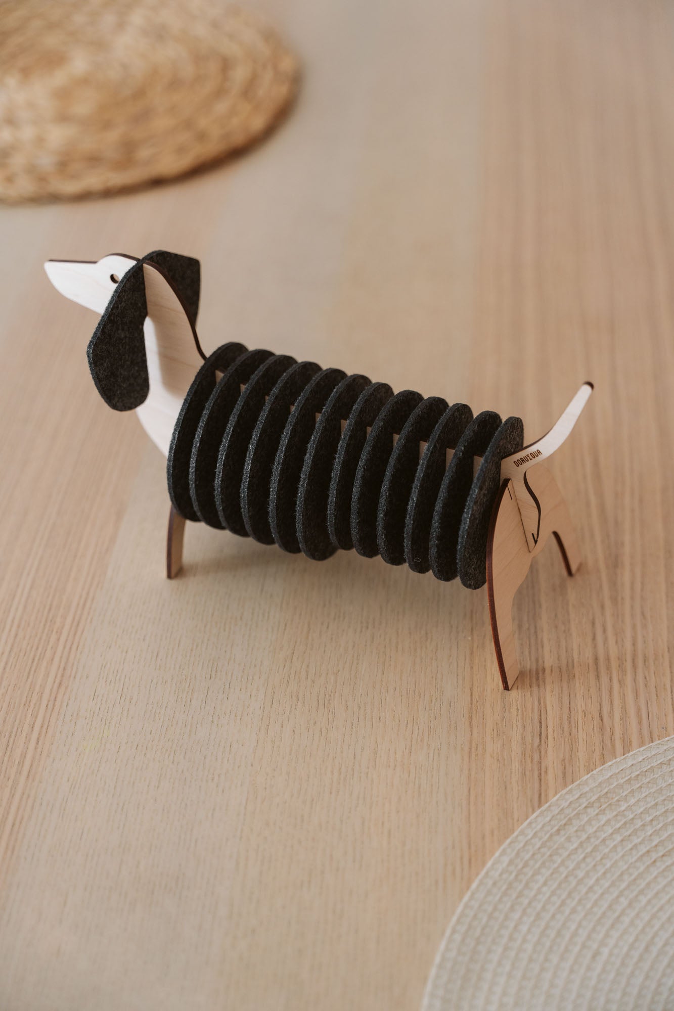 A napkin holder shaped like a dachshund dog, made of wood and black felt, sits on a light wooden table with woven placemats and matching Dachshund coasters visible in the background.
