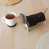 A wooden dachshund-shaped coaster holder with black round felt coasters on its body sits on a light wooden table next to a white mug, with woven placemats in the background.