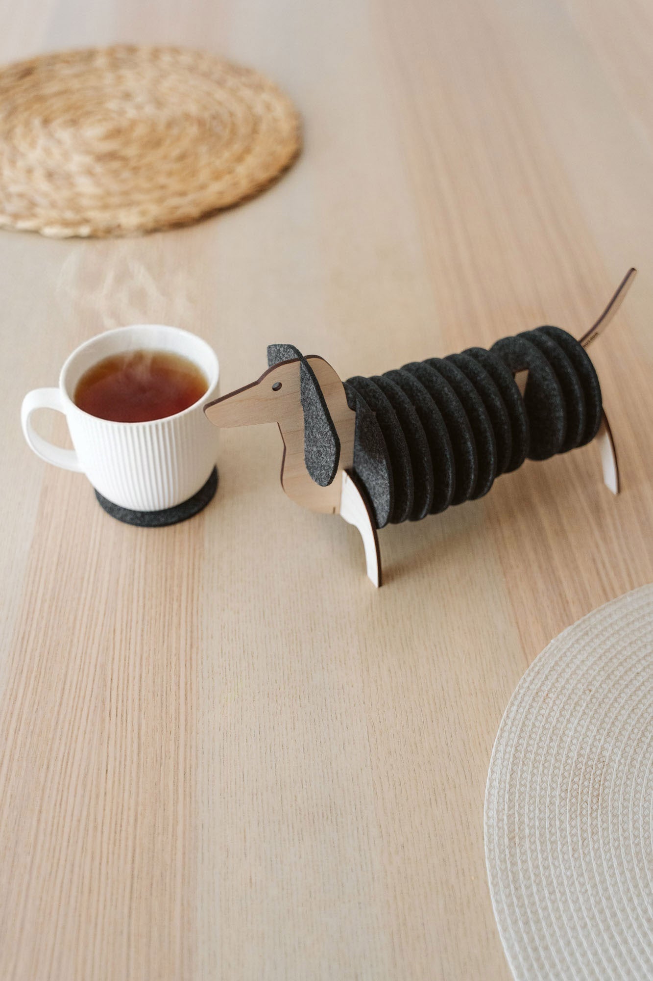 A wooden dachshund-shaped coaster holder with black round felt coasters on its body sits on a light wooden table next to a white mug, with woven placemats in the background.