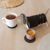 A dachshund-shaped coaster holder with black felt coasters on its body sits on a wooden table next to a white mug and a pink cup, both placed on coasters. Woven placemats are also visible on the table.