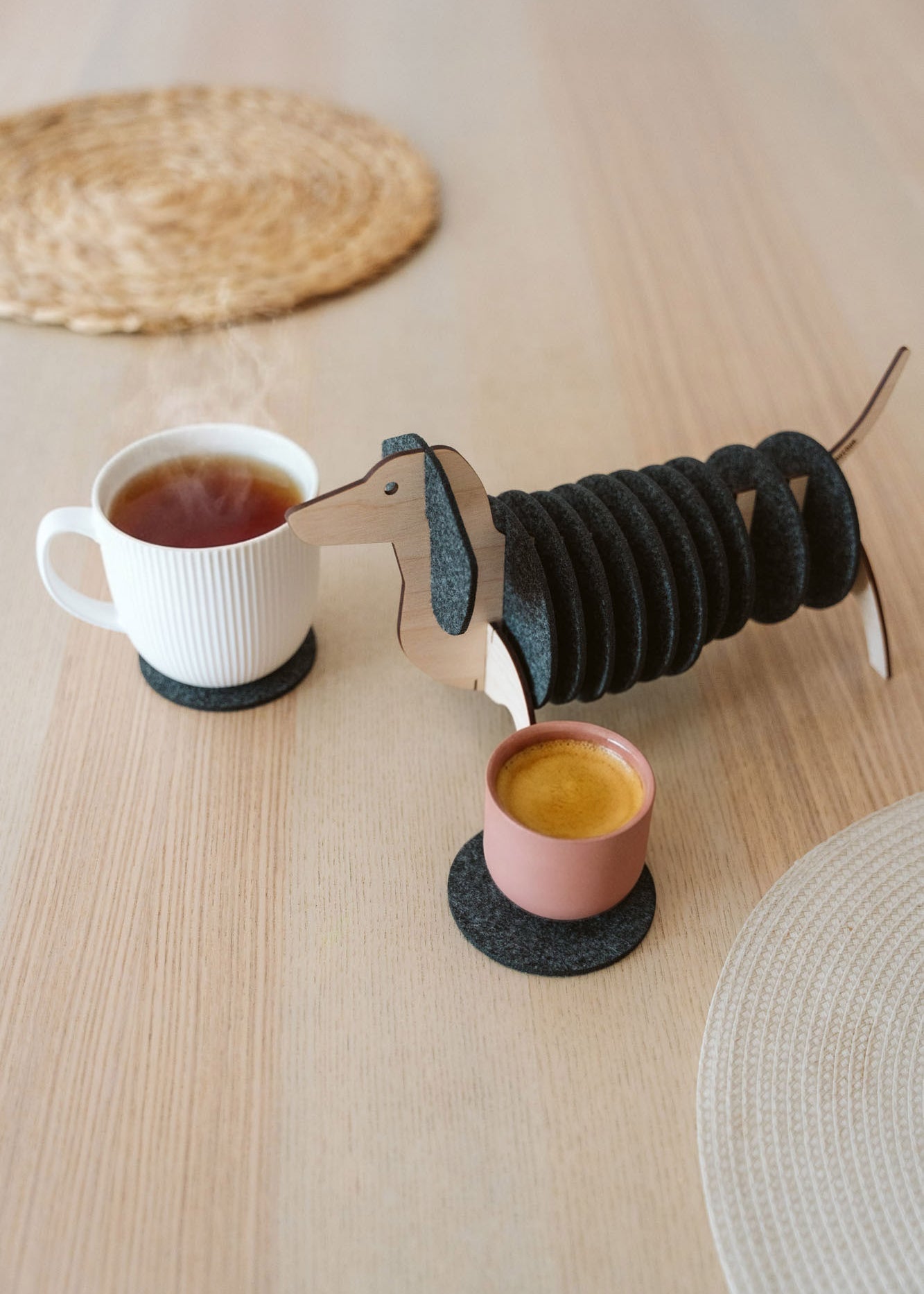 A dachshund-shaped coaster holder with black felt coasters on its body sits on a wooden table next to a white mug and a pink cup, both placed on coasters. Woven placemats are also visible on the table.