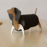 A coaster holder shaped like a dachshund dog, with a wooden head, tail, and legs, displays stacked round black felt coasters as its body, sitting on a light wood surface.