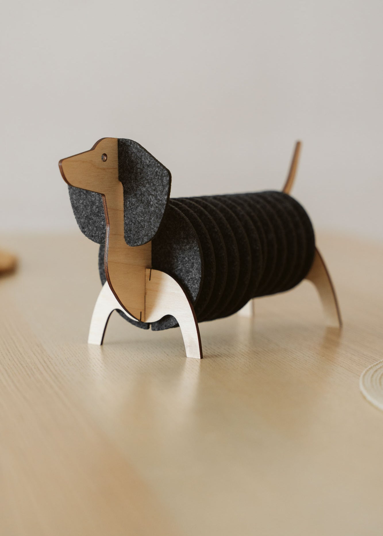 A coaster holder shaped like a dachshund dog, with a wooden head, tail, and legs, displays stacked round black felt coasters as its body, sitting on a light wood surface.