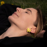 A woman with closed eyes lies on grass, smiling softly. She wears a black top and stylish Daffodil Stud Earrings—unique accessories that catch the sunlight, gently illuminating her peaceful face.