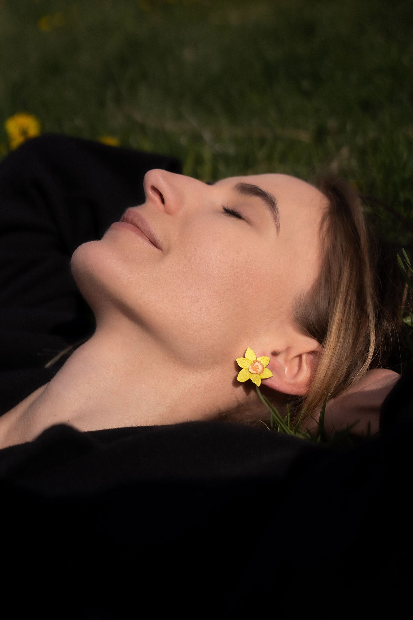 A woman with closed eyes lies on grass, smiling softly. She wears a black top and stylish Daffodil Stud Earrings—unique accessories that catch the sunlight, gently illuminating her peaceful face.