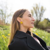A stylish woman with long, straight hair reclines on grass outdoors, her unique accessories on display—Daffodil Stud Earrings adding charm as she looks peaceful, surrounded by blurred trees and greenery.