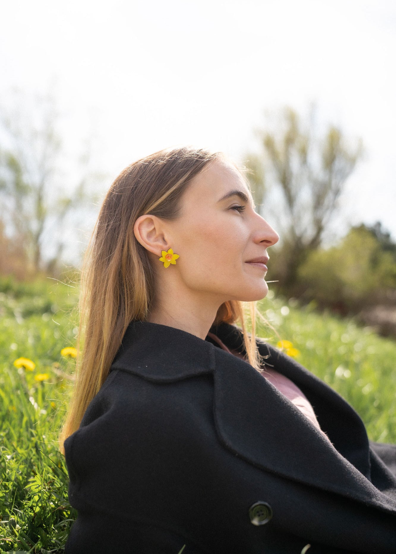 A stylish woman with long, straight hair reclines on grass outdoors, her unique accessories on display—Daffodil Stud Earrings adding charm as she looks peaceful, surrounded by blurred trees and greenery.