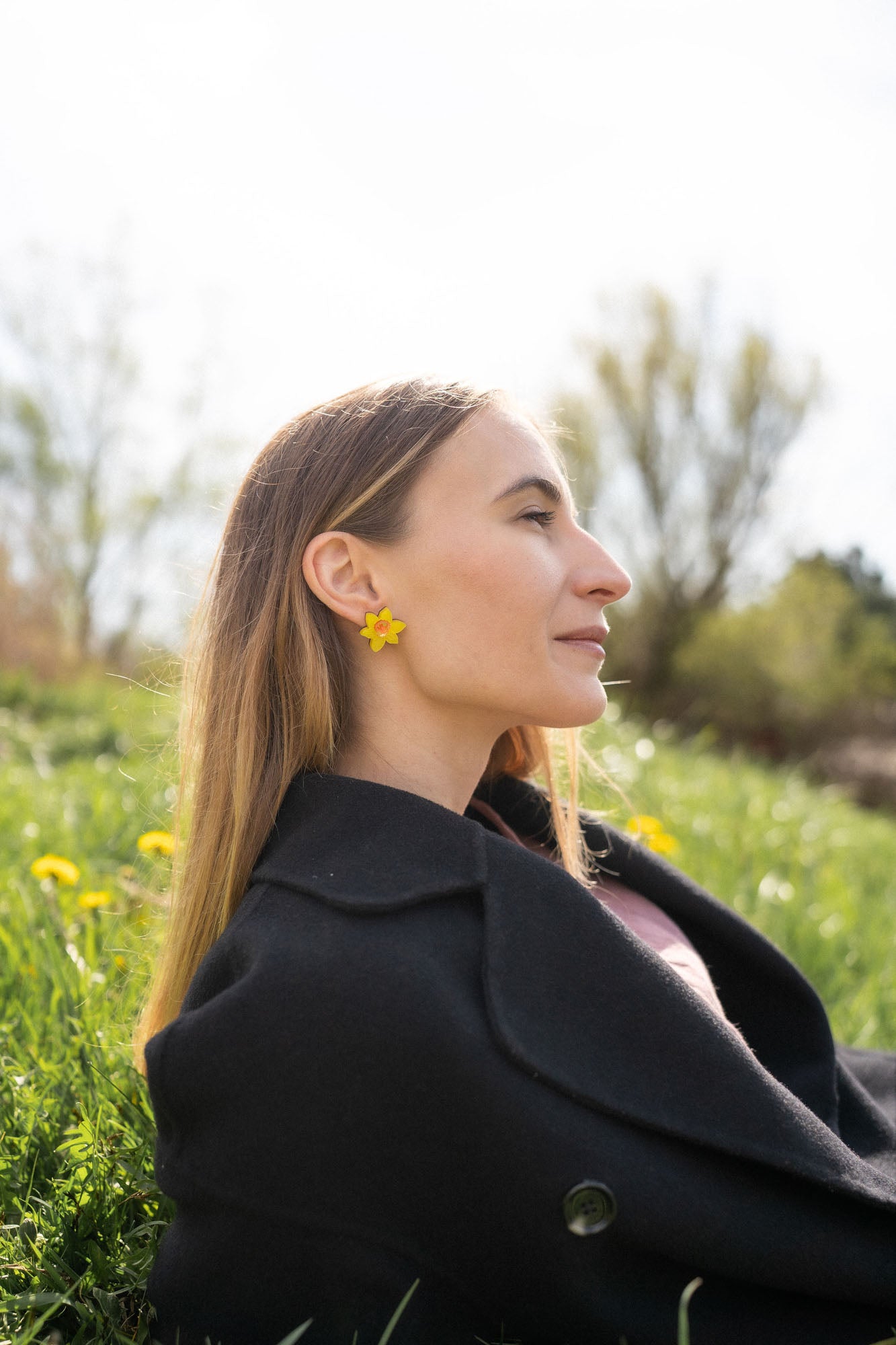 A stylish woman with long, straight hair reclines on grass outdoors, her unique accessories on display—Daffodil Stud Earrings adding charm as she looks peaceful, surrounded by blurred trees and greenery.