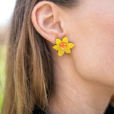 Close-up of a womans ear wearing Daffodil Stud Earrings with an orange center. Her light brown hair is tucked behind her ear, and she is dressed in a black top—perfect unique accessories for stylish women.