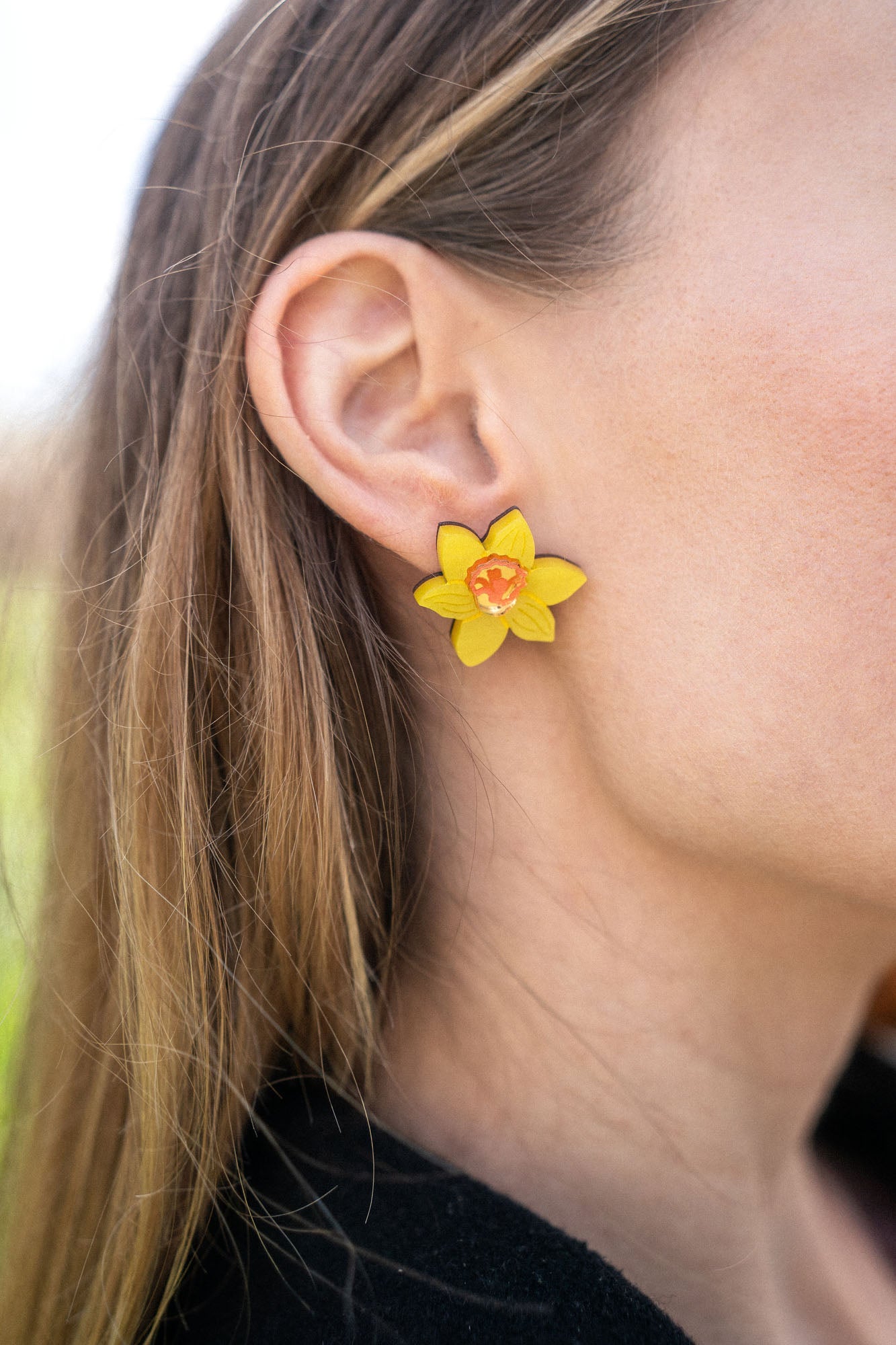 Close-up of a womans ear wearing Daffodil Stud Earrings with an orange center. Her light brown hair is tucked behind her ear, and she is dressed in a black top—perfect unique accessories for stylish women.