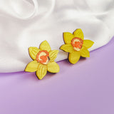 Two yellow Daffodil Stud Earrings with orange centers rest on soft white fabric, set against a pastel purple background—unique accessories perfect for stylish women seeking a floral touch.
