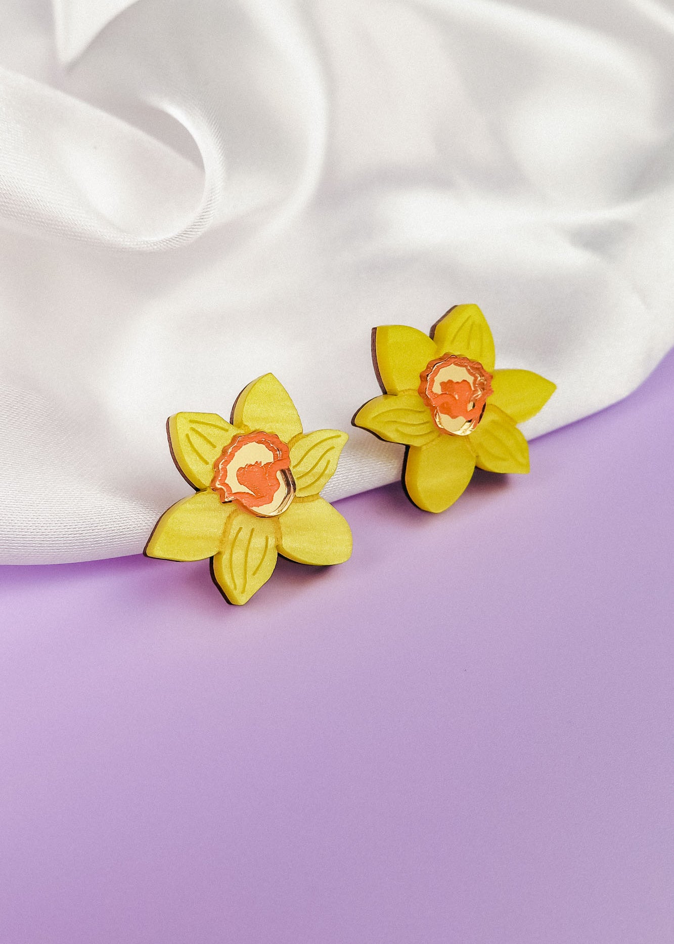 Two yellow Daffodil Stud Earrings with orange centers rest on soft white fabric, set against a pastel purple background—unique accessories perfect for stylish women seeking a floral touch.