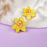 Two yellow daffodil stud earrings, unique accessories for stylish women, rest on soft white fabric and a purple surface, with a blurred beige element in the foreground.