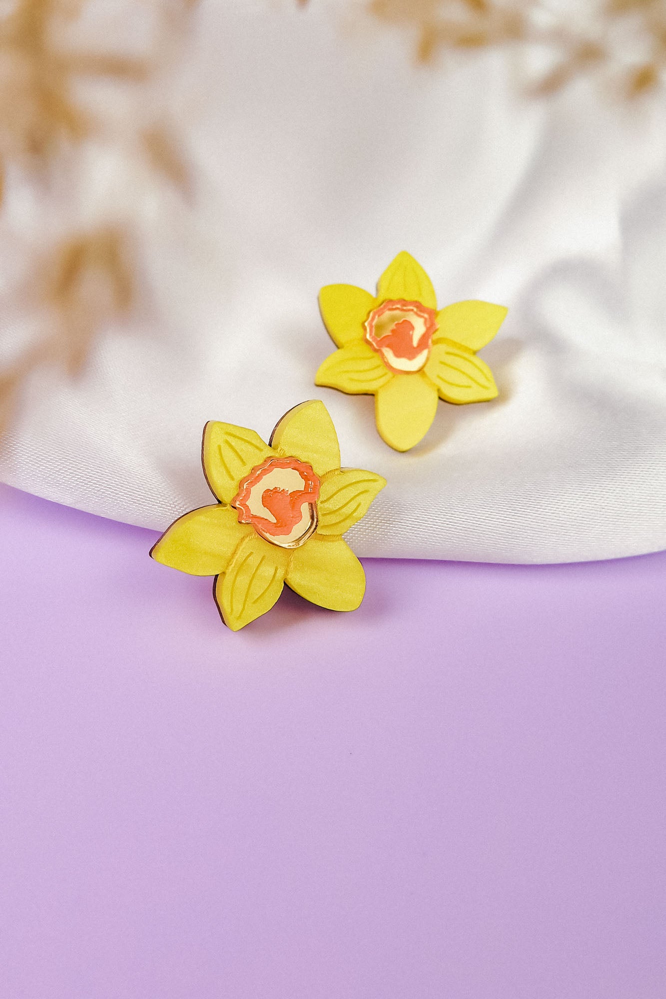 Two yellow daffodil stud earrings, unique accessories for stylish women, rest on soft white fabric and a purple surface, with a blurred beige element in the foreground.