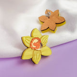 A pair of Daffodil Stud Earrings for stylish women—one features a yellow daffodil with a red center, while the other reveals the plain wooden back. These unique accessories are displayed on white fabric and a purple surface.