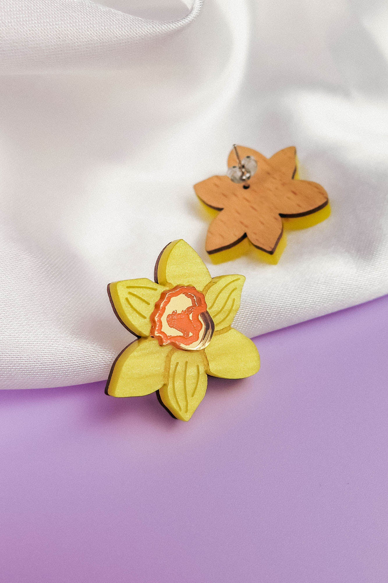 A pair of Daffodil Stud Earrings for stylish women—one features a yellow daffodil with a red center, while the other reveals the plain wooden back. These unique accessories are displayed on white fabric and a purple surface.