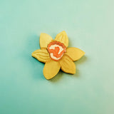 A handmade yellow flower-shaped cookie with an orange and white center is placed on a light teal background, reminiscent of a Daffodil Flower Lapel Pin crafted in acrylic.