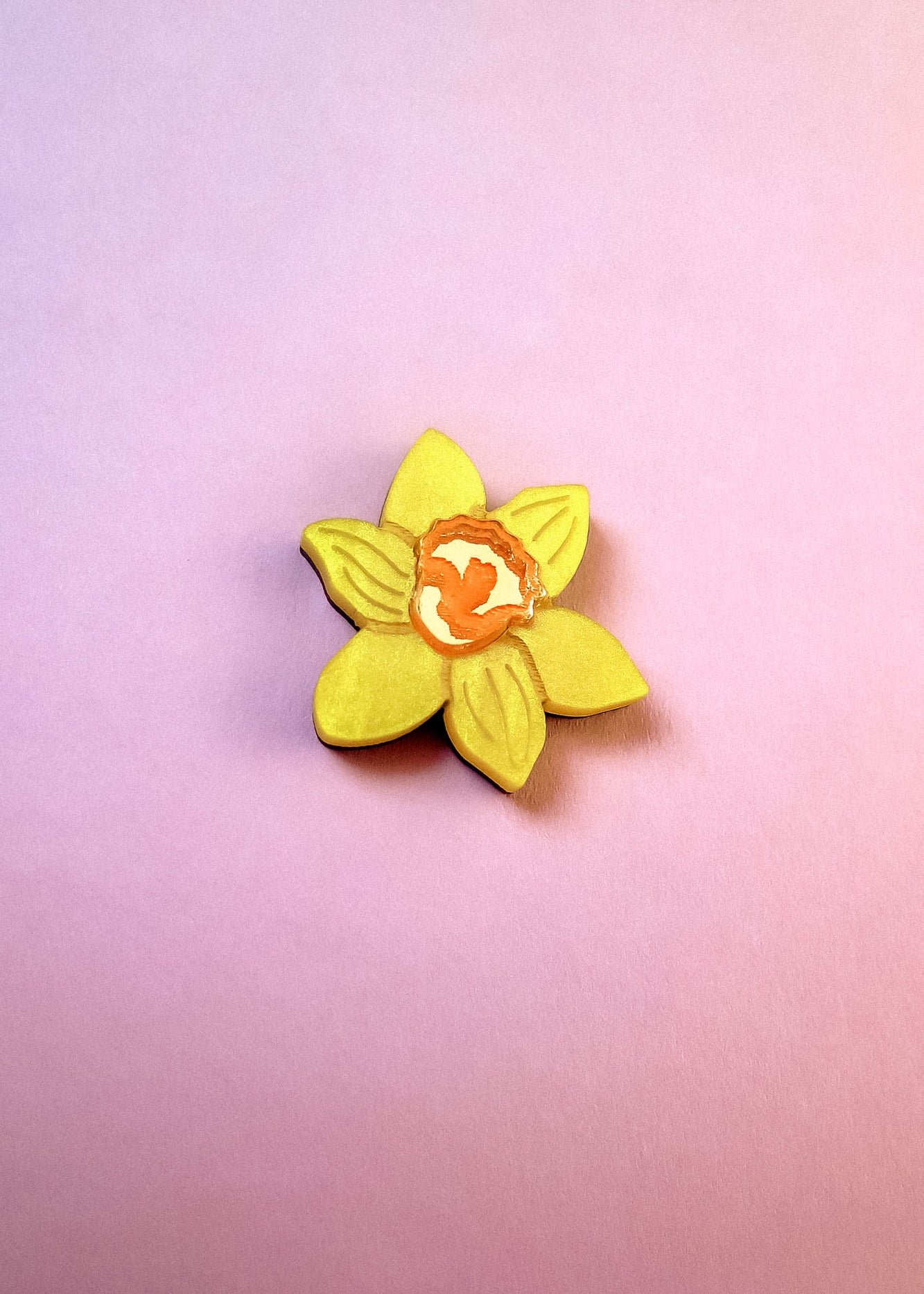A handmade yellow Daffodil Flower Lapel Pin with an orange center is placed on a pink and purple gradient background.