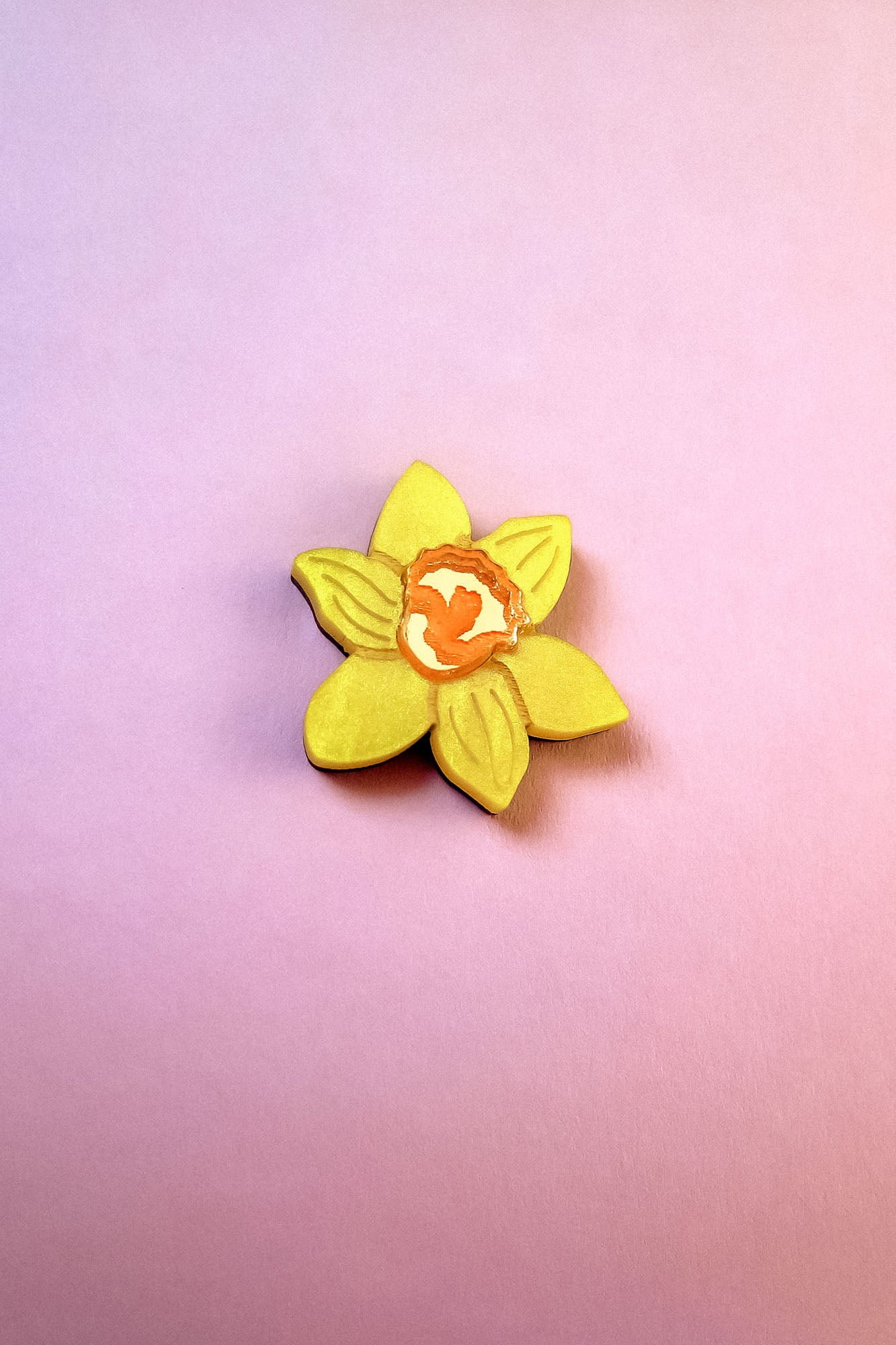 A handmade yellow Daffodil Flower Lapel Pin with an orange center is placed on a pink and purple gradient background.
