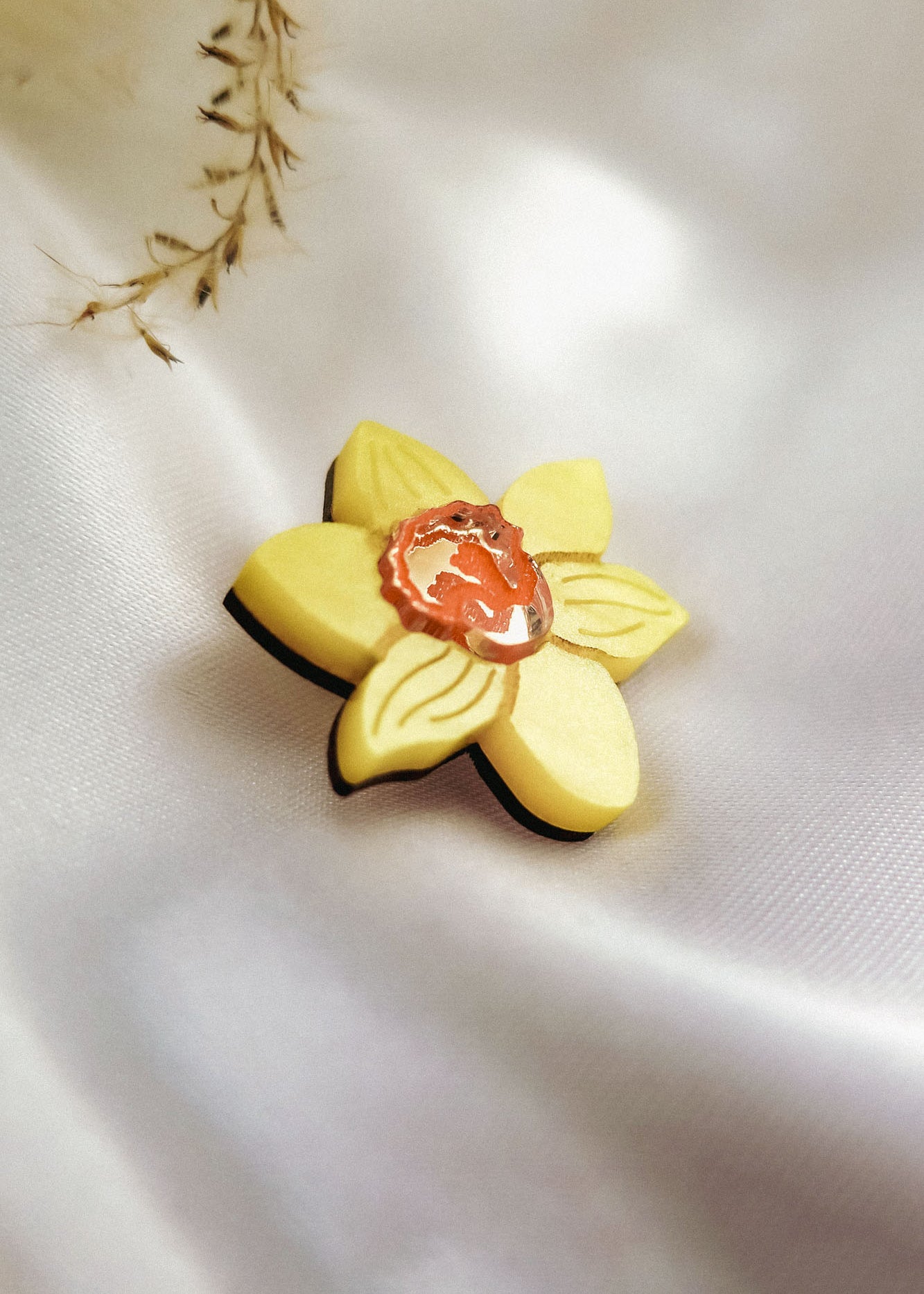 A handmade yellow Daffodil Flower Lapel Pin brooch with a small orange and white cameo in the center, crafted from acrylic, rests on soft white satin fabric. Delicate dried plant stems add a subtle touch in the background.