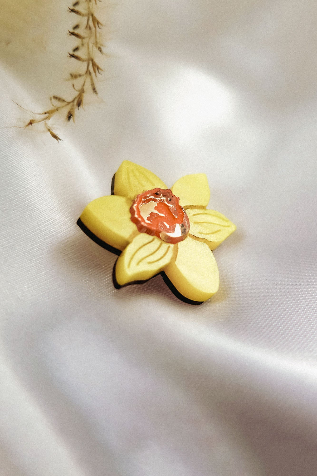 A handmade yellow Daffodil Flower Lapel Pin brooch with a small orange and white cameo in the center, crafted from acrylic, rests on soft white satin fabric. Delicate dried plant stems add a subtle touch in the background.