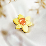 A yellow, handmade flower-shaped cookie with an orange center rests on white, silky fabric, resembling a Daffodil Flower Lapel Pin, with blurred brownish plants in the background.