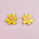Two yellow Daffodil Stud Earrings with orange centers, resembling, are arranged side by side on a light pink background—perfect for stylish women seeking unique accessories.
