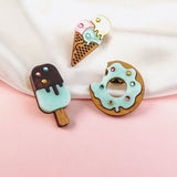 Three wooden pins shaped like desserts—a popsicle, an ice cream cone, and a handcrafted Donut Pin—decorated with pastel icing and colorful sprinkles, are displayed on a pink and white background.