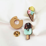 Three handcrafted enamel pins on white fabric: a Donut Pin with a bite taken out, a triple scoop ice cream cone, and a chocolate-covered popsicle, each sparkling with rhinestones and paired with a gold pin backing nearby.