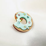 A handcrafted wooden Donut Pin shaped like a bitten donut, topped with pale blue icing and colorful sprinkles, rests on a soft white fabric background.