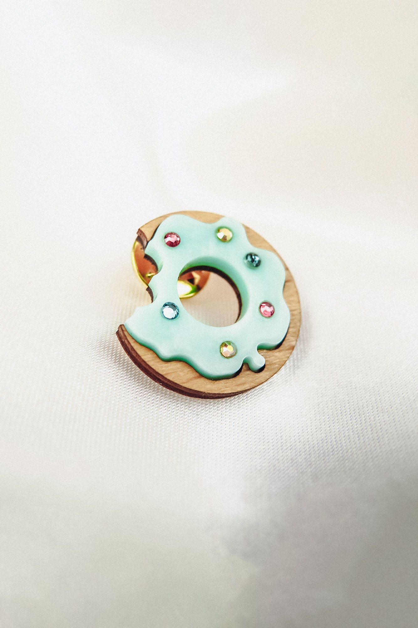 A handcrafted wooden Donut Pin shaped like a bitten donut, topped with pale blue icing and colorful sprinkles, rests on a soft white fabric background.