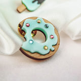 A handcrafted wooden Donut Pin with light blue icing and sparkling rhinestones rests on white fabric. In the background, blurred pins shaped like an ice cream cone and a popsicle add a playful touch.