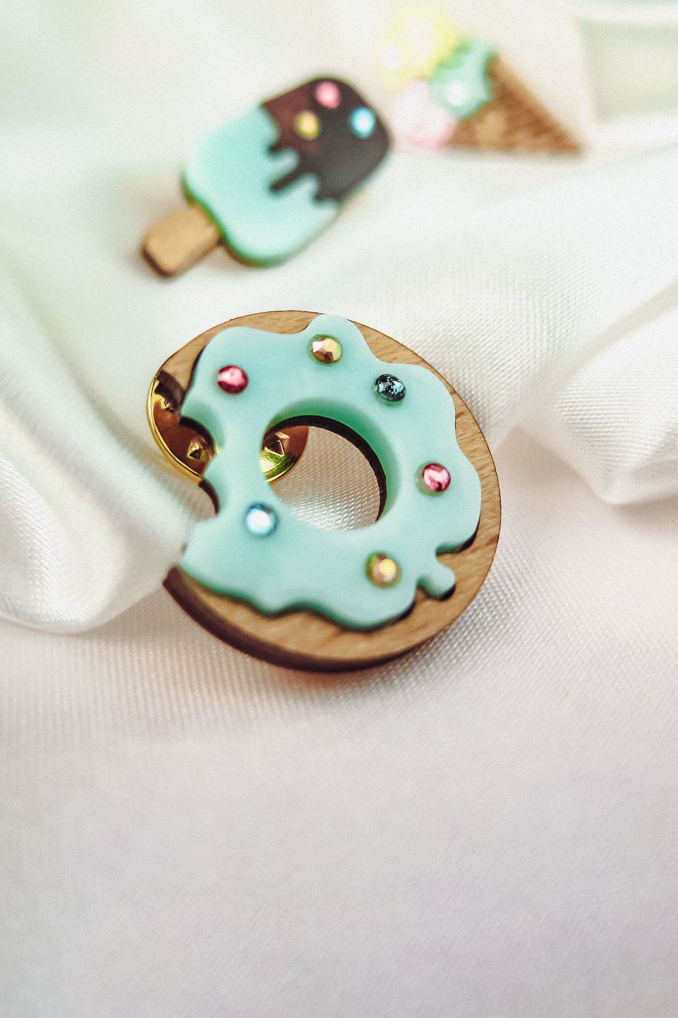 A handcrafted wooden Donut Pin with light blue icing and sparkling rhinestones rests on white fabric. In the background, blurred pins shaped like an ice cream cone and a popsicle add a playful touch.