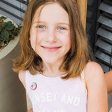 A young girl with shoulder-length light brown hair and blue eyes smiles at the camera. She wears a pale pink sleeveless top reading SUNSET AND PALMS, paired with a handcrafted Donut Pin. Plants and wooden details decorate the background.