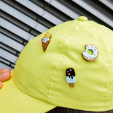 A person wears a bright yellow cap adorned with three cute handcrafted pins: an ice cream cone, a donut pin sparkling with rhinestones and sprinkles, and a chocolate-covered popsicle. Venetian blinds are in the background.