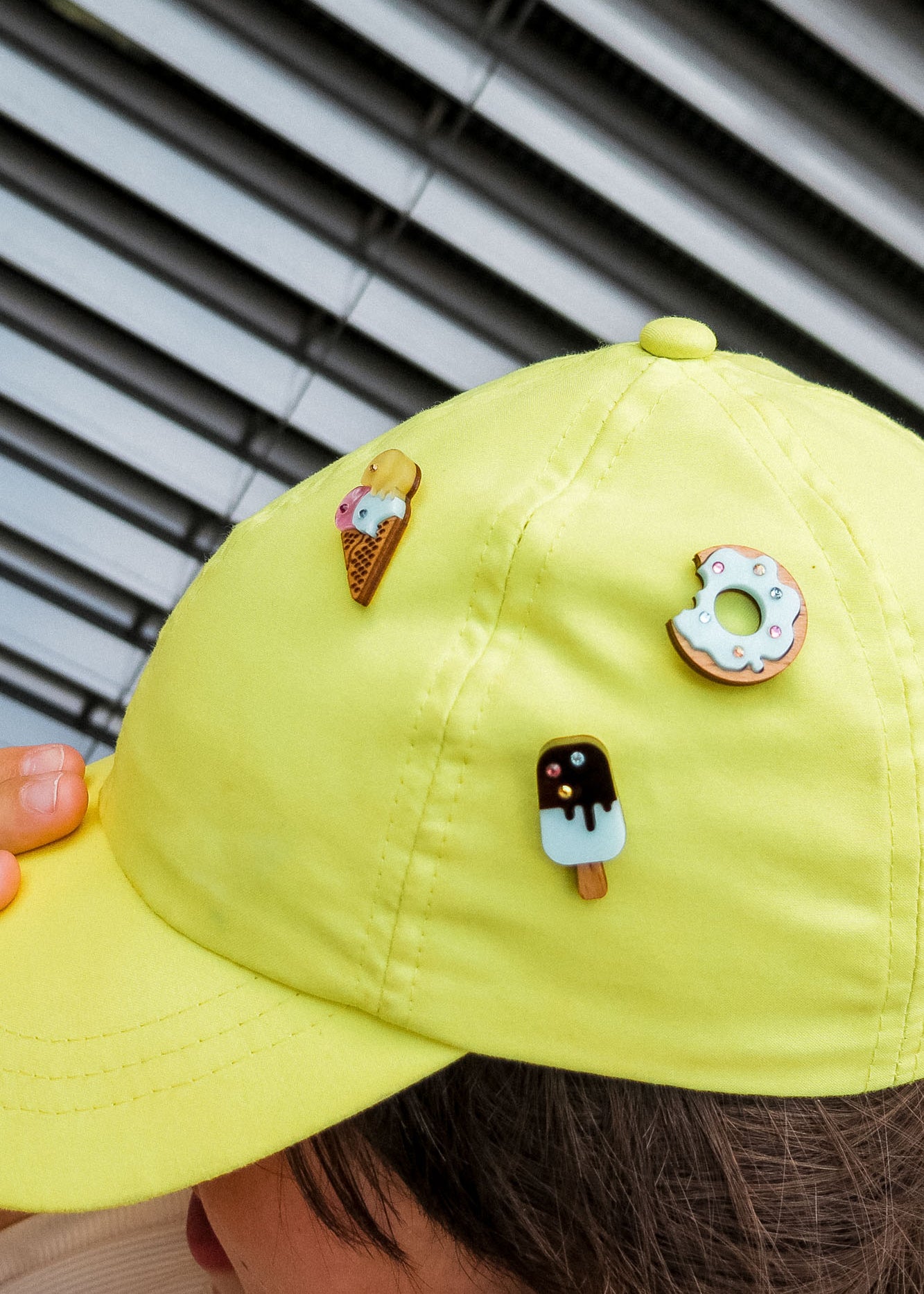 A person wears a bright yellow cap adorned with three cute handcrafted pins: an ice cream cone, a donut pin sparkling with rhinestones and sprinkles, and a chocolate-covered popsicle. Venetian blinds are in the background.