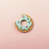 A handcrafted Donut Pin featuring a wooden base, light blue icing, gem-like rhinestones, and a bite taken out, set against a pastel pink background.