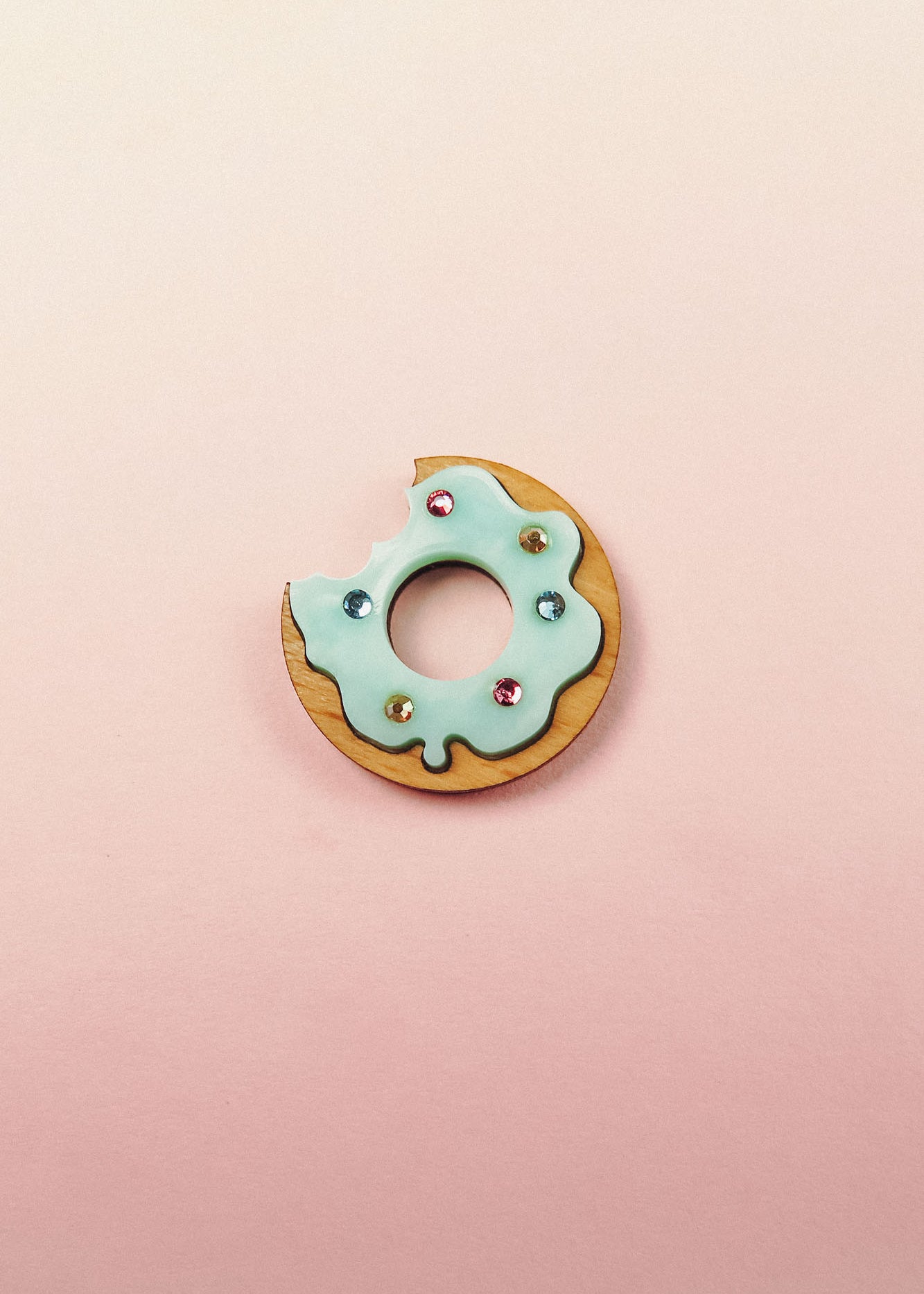 A handcrafted Donut Pin featuring a wooden base, light blue icing, gem-like rhinestones, and a bite taken out, set against a pastel pink background.
