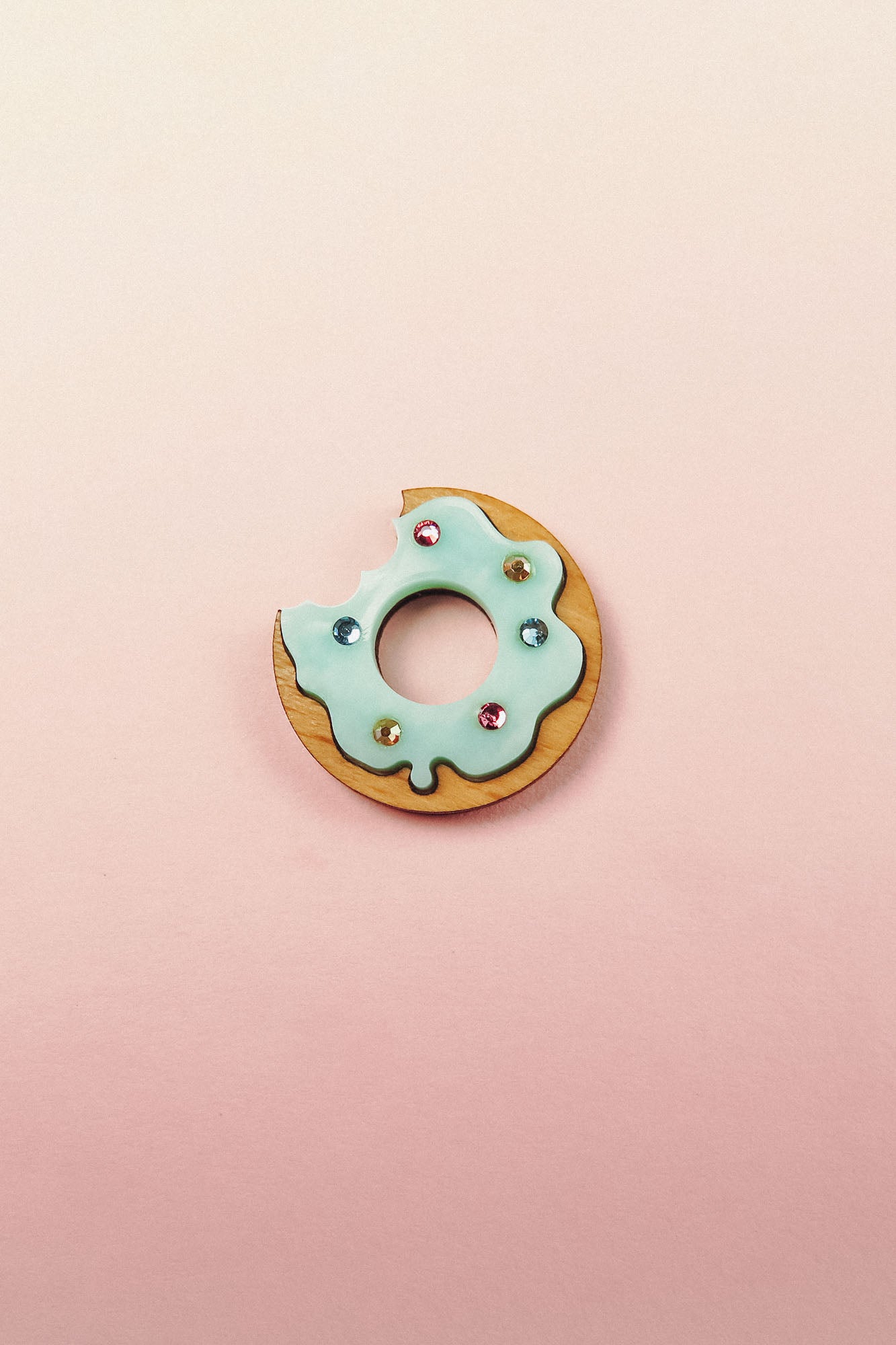 A handcrafted Donut Pin featuring a wooden base, light blue icing, gem-like rhinestones, and a bite taken out, set against a pastel pink background.