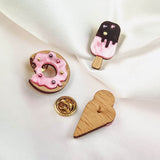 Three handcrafted wooden pins on white fabric: a pink frosted Donut Pin with sprinkles, a pink and brown ice cream pop adorned with colorful rhinestones, and a plain wooden heart. One gold pin backing is also visible.