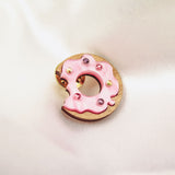 A pink frosted donut-shaped pin with colorful rhinestones and a bite taken out of it rests on soft, white satin fabric.