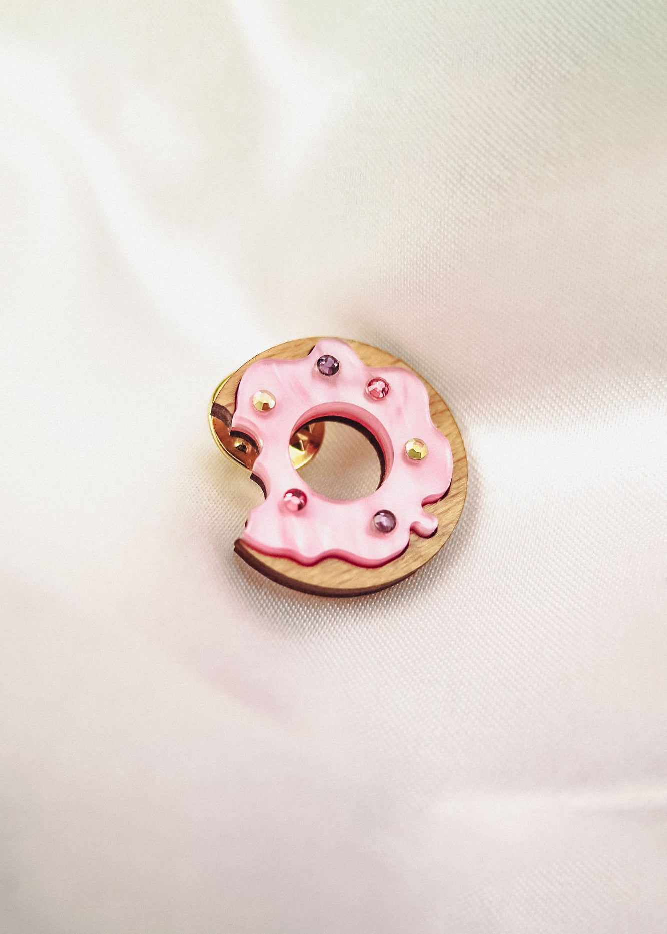 A pink frosted donut-shaped pin with colorful rhinestones and a bite taken out of it rests on soft, white satin fabric.