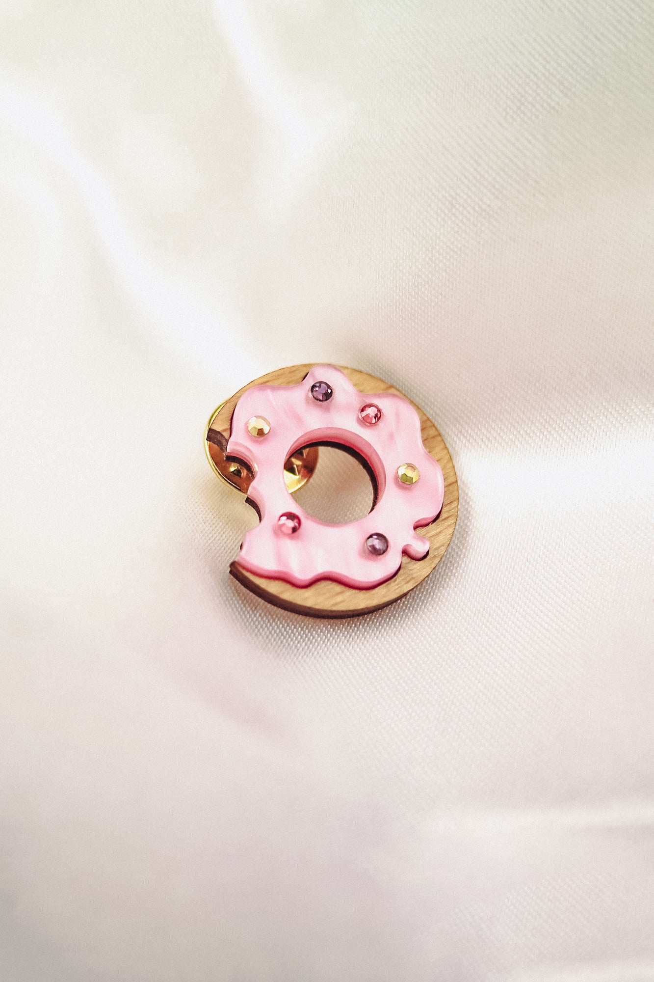 A pink frosted donut-shaped pin with colorful rhinestones and a bite taken out of it rests on soft, white satin fabric.