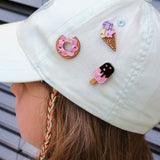 A child wearing a light-colored cap decorated with three cute handcrafted pins—a pink Donut Pin, an ice cream cone, and a popsicle—each adorned with colorful rhinestones. The background features horizontal gray metal slats.