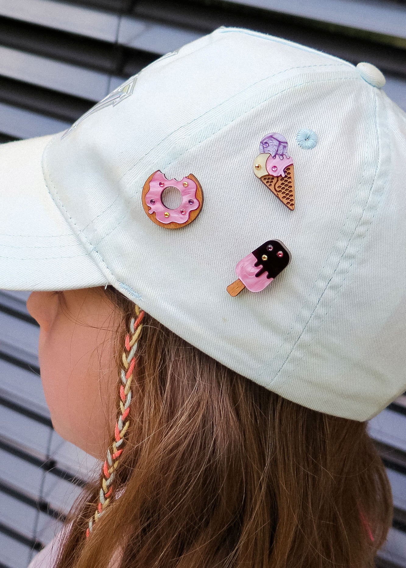 A child wearing a light-colored cap decorated with three cute handcrafted pins—a pink Donut Pin, an ice cream cone, and a popsicle—each adorned with colorful rhinestones. The background features horizontal gray metal slats.